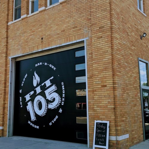Exterior of a brick building featuring a large black garage door with "Pit 105" logo and a sign indicating lunch hours.