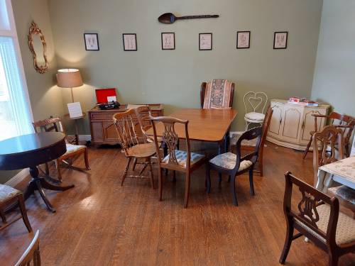 A cozy room with wooden floors, various chairs, a table, and framed artwork on the walls. A lamp and vintage decor are present.