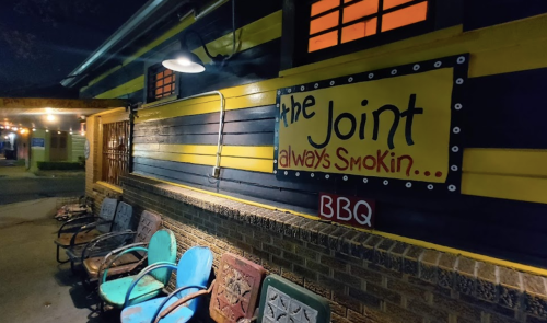 A brightly painted building with a sign reading "the Joint always Smokin... BBQ" and colorful chairs outside.
