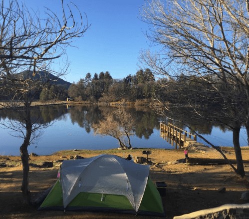 A tent by a calm lake surrounded by trees, with a dock and mountains in the background under a clear blue sky.