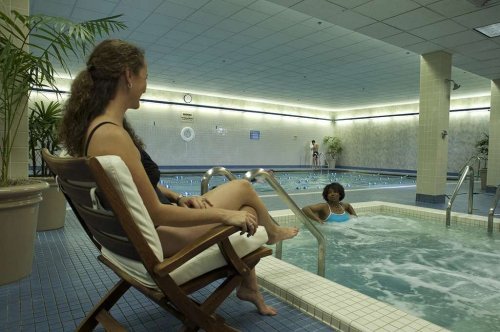 Two women relax in a hotel pool area; one sits on a lounge chair while the other enjoys the water.