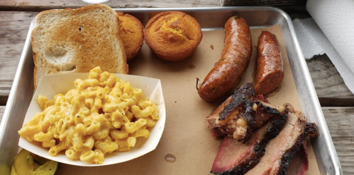A tray with macaroni and cheese, sausage links, brisket slices, cornbread muffins, and toasted bread.