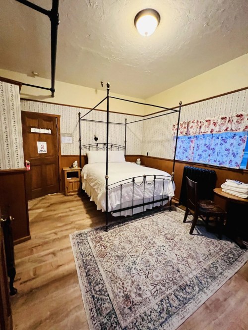 Cozy bedroom with a four-poster bed, wooden furniture, and floral wallpaper, featuring a rug and window with curtains.