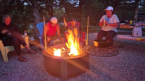 A group of four people sitting around a campfire, enjoying drinks and snacks in a cozy outdoor setting.