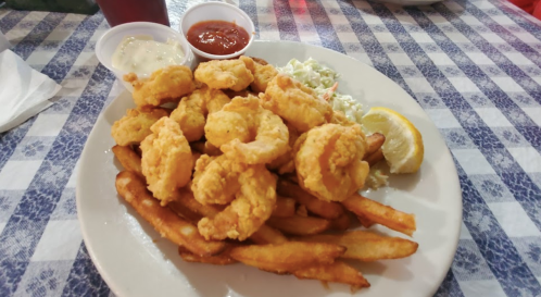 A plate of golden fried shrimp served with fries, coleslaw, lemon, and dipping sauces on a checkered tablecloth.
