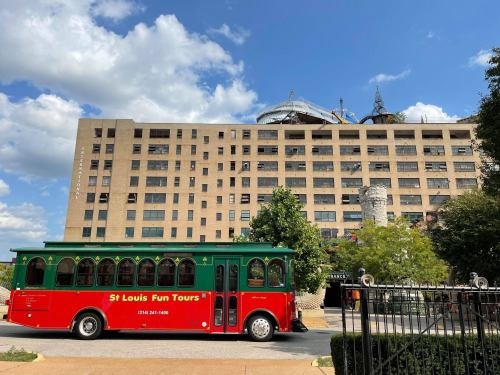 A red and green trolley bus parked in front of a large, historic building under a partly cloudy sky.