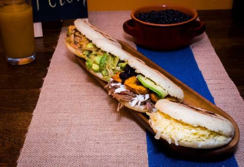 A long wooden platter holds a stuffed sandwich with various toppings, alongside a bowl of black beans and a drink.