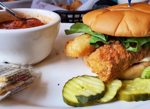 A crispy fish sandwich with lettuce, pickles, and a side of marinara sauce in a bowl.