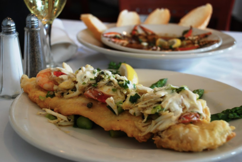 A plate of fried fish topped with shrimp, crab, and vegetables, served with lemon and a side of bread.
