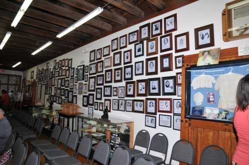 A wall filled with framed black-and-white photos and display cases in a museum setting, with chairs in the foreground.