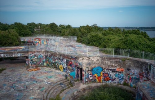 Abandoned concrete structure covered in colorful graffiti, surrounded by lush greenery and overlooking a body of water.