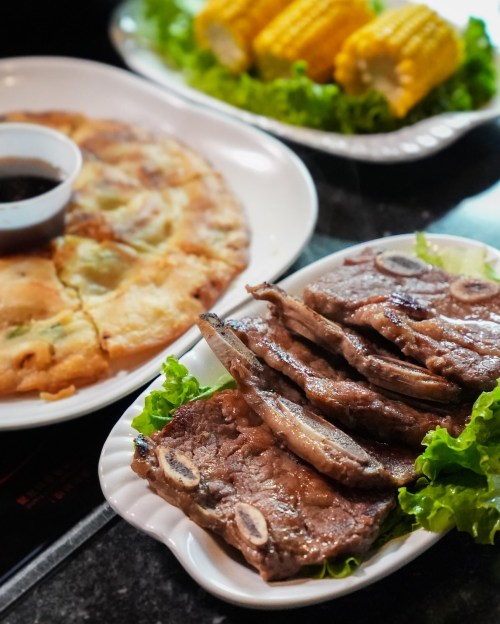 A plate of grilled ribs on lettuce, accompanied by a dipping sauce, corn on the cob, and a savory pancake.