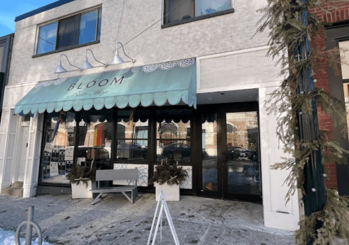 A storefront with a light blue awning reading "BLOOM" and snow on the ground, featuring potted plants by the entrance.
