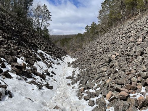 A rocky path between two steep slopes, with patches of snow and trees in the background under a cloudy sky.