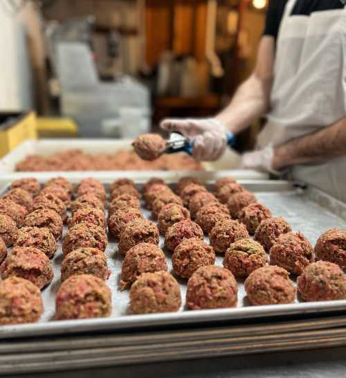 A chef scoops meatballs onto a tray, surrounded by rows of uncooked meatballs in a kitchen setting.