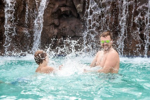 A man and a child play in a pool, splashing water with a waterfall in the background. Both are smiling and enjoying.
