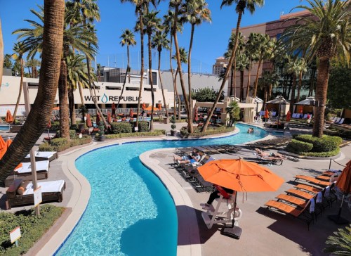 A sunny pool area with a winding blue pool, orange umbrellas, and palm trees, surrounded by lounge chairs.