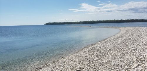 A serene shoreline with smooth pebbles, clear water, and a distant tree-lined island under a blue sky.