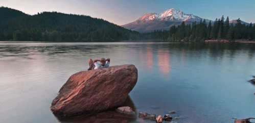 A person relaxes on a large rock by a serene lake, surrounded by mountains and lush trees at sunset.