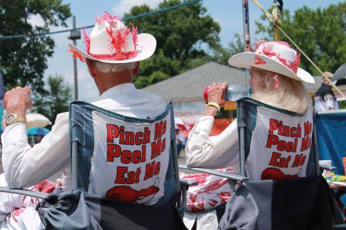 Two elderly individuals in matching hats and shirts with playful lobster-themed slogans, sitting in folding chairs at an outdoor event.