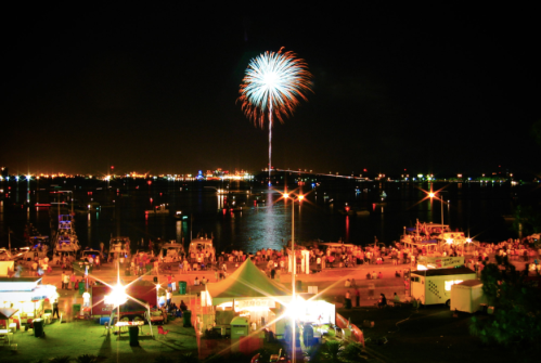 Fireworks illuminate the night sky over a bustling waterfront, with boats and crowds enjoying the festive atmosphere.
