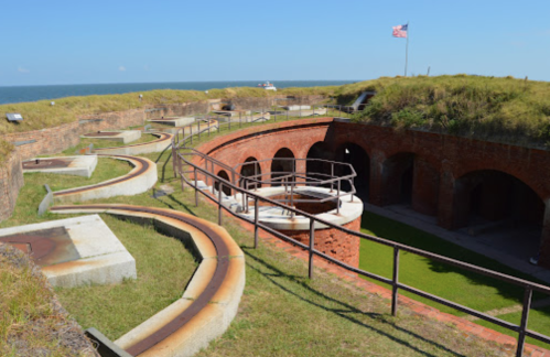 Historic fort with curved pathways, grassy areas, and a flag, overlooking the ocean under a clear blue sky.