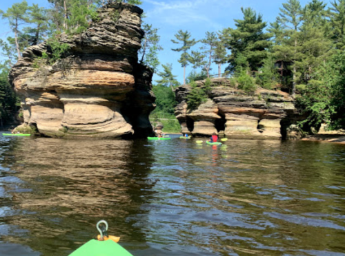 Kayakers paddle near large rock formations surrounded by trees and a clear blue sky.
