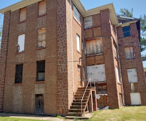 Abandoned brick building with boarded windows and a staircase, set against a clear blue sky.