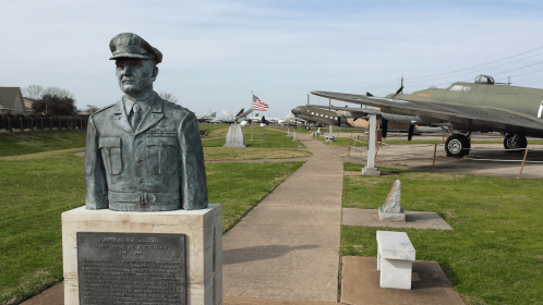 A bronze bust of a military officer stands in a park with vintage aircraft and an American flag in the background.