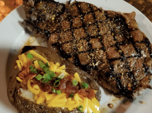 A grilled steak with a crisscross pattern, served alongside a loaded baked potato topped with cheese, bacon, and green onions.
