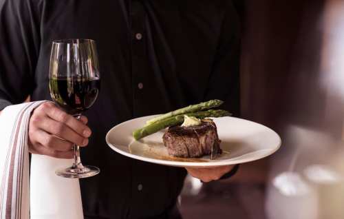 A person holding a plate with a steak and asparagus, and a glass of red wine in the other hand.