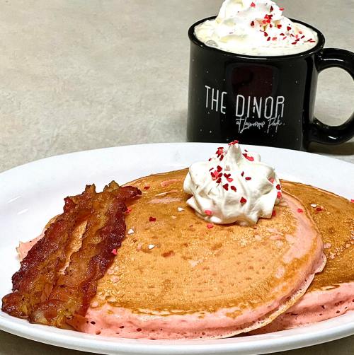 A plate of pink pancakes topped with whipped cream and bacon, alongside a mug of whipped cream-topped hot chocolate.
