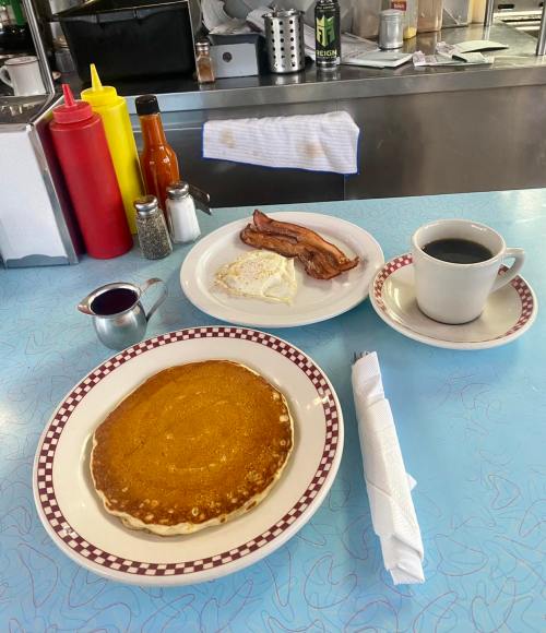 A breakfast plate with pancakes, bacon, eggs, and a cup of coffee on a blue countertop. Condiments in the background.