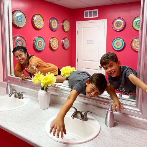 A woman and two children playfully pose in a colorful bathroom with flowers and circular wall decor.