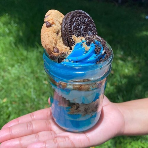 A hand holds a jar filled with blue frosting, cookie crumbles, and topped with cookies and an Oreo.