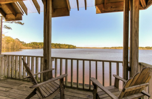 View from a wooden porch overlooking a calm lake surrounded by trees under a clear blue sky.