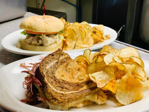A plate with a grilled sandwich and crispy potato chips, alongside a burger with lettuce and tomato on a bun.