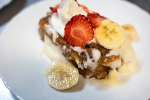 A slice of dessert topped with whipped cream, strawberries, and banana slices, drizzled with creamy sauce on a white plate.