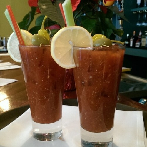 Two tall glasses of Bloody Mary cocktails garnished with lemon slices, olives, and celery, set on a bar counter.