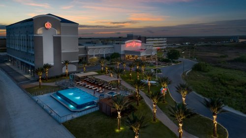 Aerial view of a hotel with a pool and palm trees at sunset, showcasing a vibrant sky and well-lit surroundings.