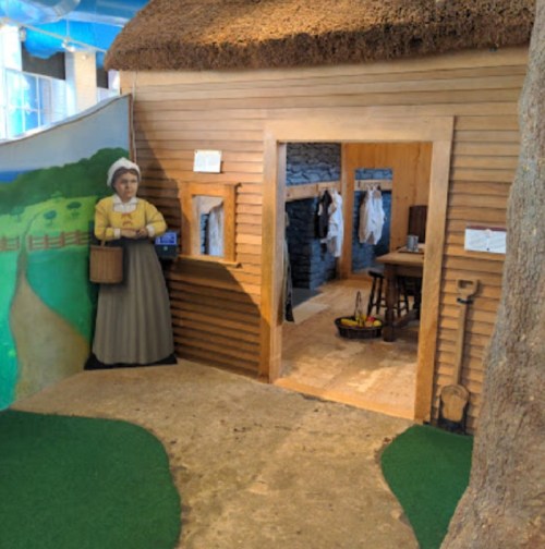 A rustic wooden house with a thatched roof, featuring a woman figure outside and a path leading to the entrance.
