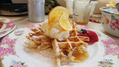 A plate of waffles topped with a poached egg, lemon slices, syrup, and a strawberry, with a floral border.