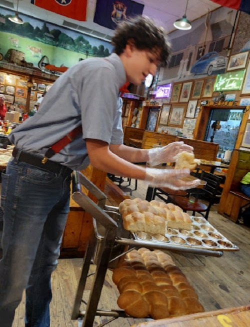A young person in a blue shirt and suspenders places rolls on a tray in a rustic restaurant filled with decor.