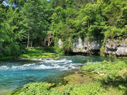 A serene river flows past lush greenery and rocky cliffs, with an old stone structure partially hidden among the trees.