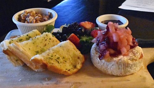 A wooden board with garlic bread, fresh fruit, nuts, and a round cheese topped with fruit compote.