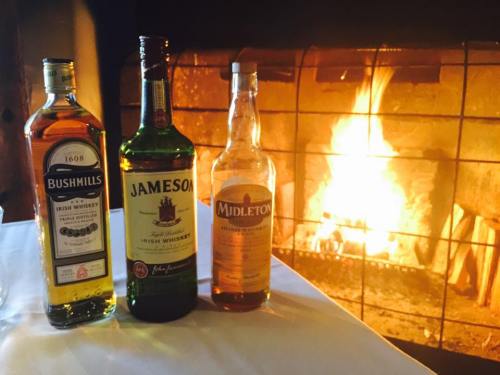 Three bottles of Irish whiskey—Bushmills, Jameson, and Midleton—set against a warm, glowing fireplace.