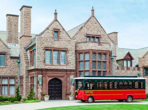 A large stone mansion with a red and green trolley parked in front, surrounded by well-maintained landscaping.