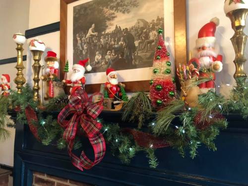 A festive mantel decorated with Santa figures, a Christmas tree, candles, and garlands, featuring a classic painting above.