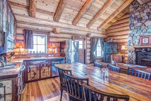 Cozy log cabin kitchen with wooden beams, a large dining table, stone fireplace, and modern appliances.