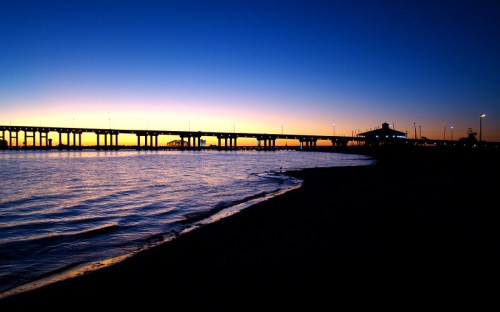 Silhouette of a pier at sunset, with calm water and a gradient sky transitioning from blue to orange.